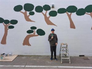 Young man standing in front of partially painted outdoor mural featuring stylised trees